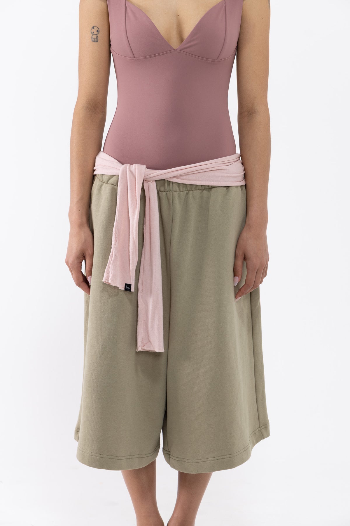 A dancer wearing a dusty rose bodysuit, olive green wide-leg shorts, and a light pink fabric belt tied at the waist, shot against a plain white studio background.