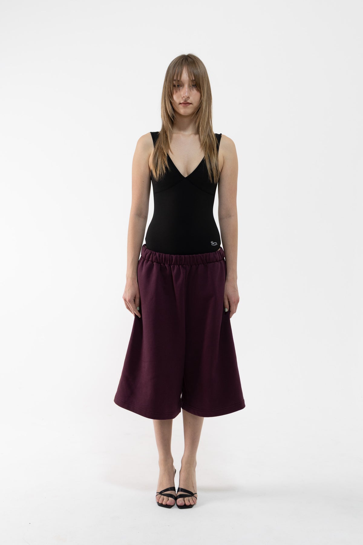 A dancer stands against a plain white studio background wearing a black sleeveless V-neck top and dark plum wide-leg shorts or culottes with black sandals.