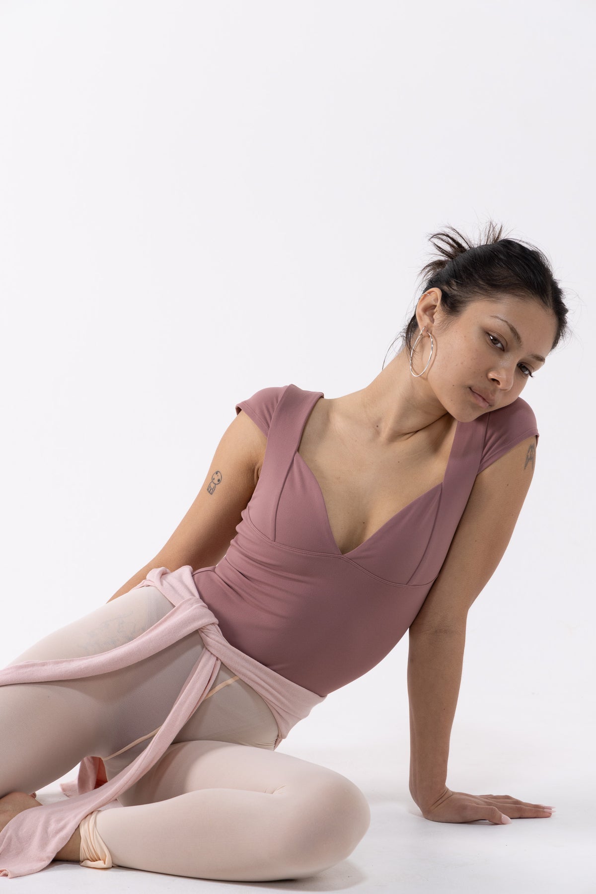 A dancer in a dusty rose dance leotard and light pink tights poses on a seamless white studio background.