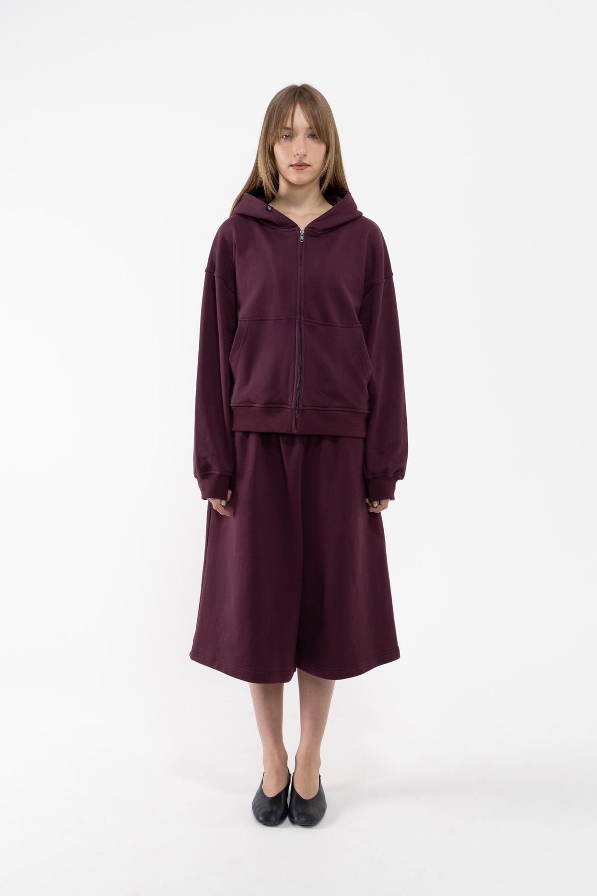 A dancer stands against a plain white studio background wearing a matching plum zip-up hoodie and wide-leg cropped sweatpants, paired with black flats.