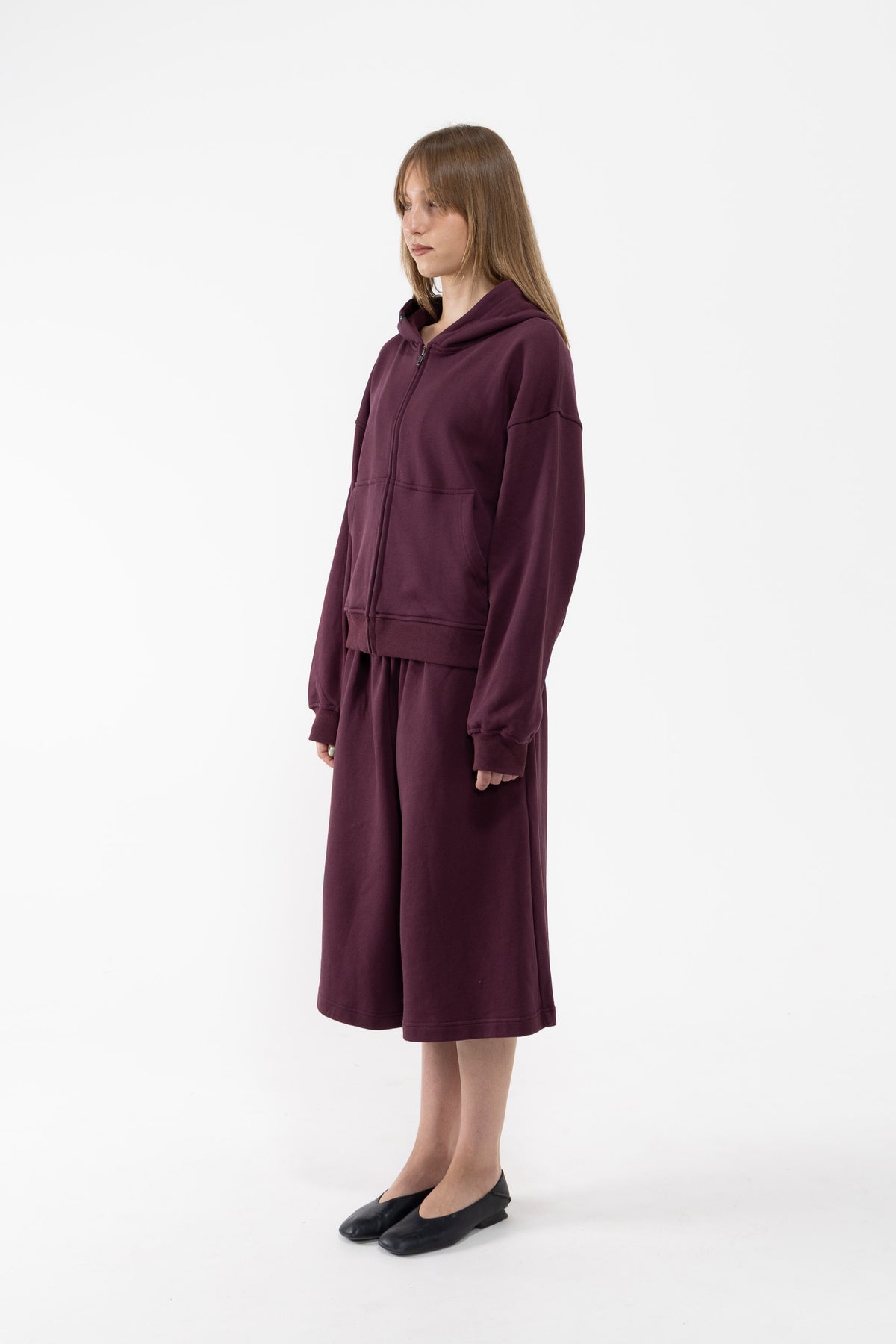 A dancer modeling a matching deep plum lounge set consisting of a zip-up hoodie and wide-leg sweatpants, paired with black flats, against a plain white studio background.