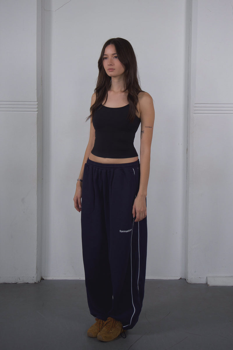 Woman wearing a black crop top and navy pants against a white wall.