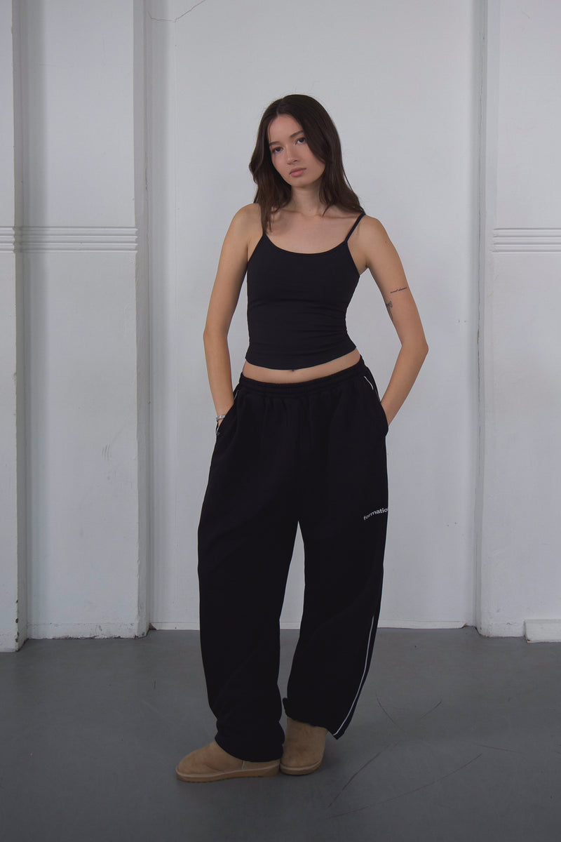 Woman wearing a black tank top and black pants standing against a white wall.