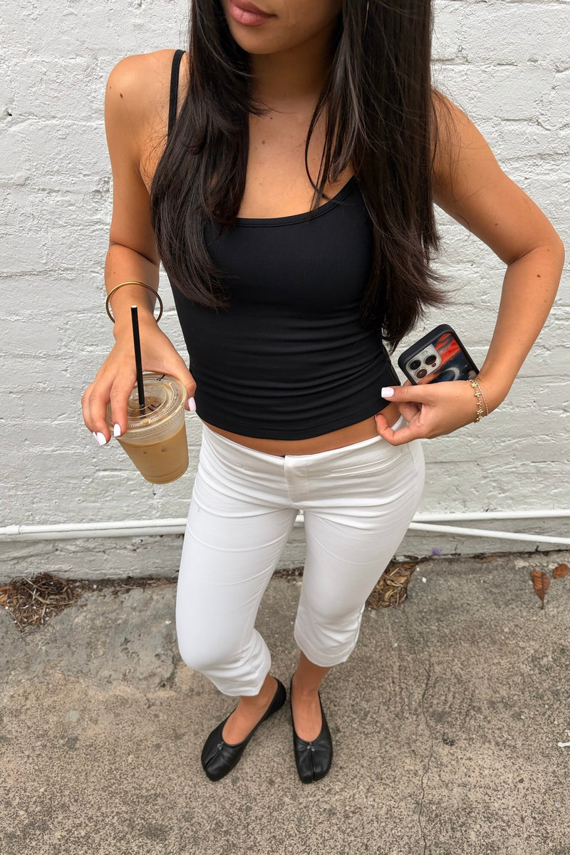 Woman in black top and white pants holding a drink and phone against a white brick wall.
