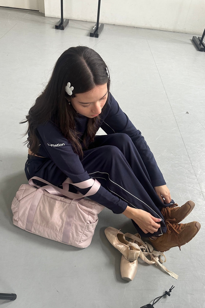 Woman sitting on the floor tying ballet shoes with a pink bag beside her. Wearing navy sweat set.