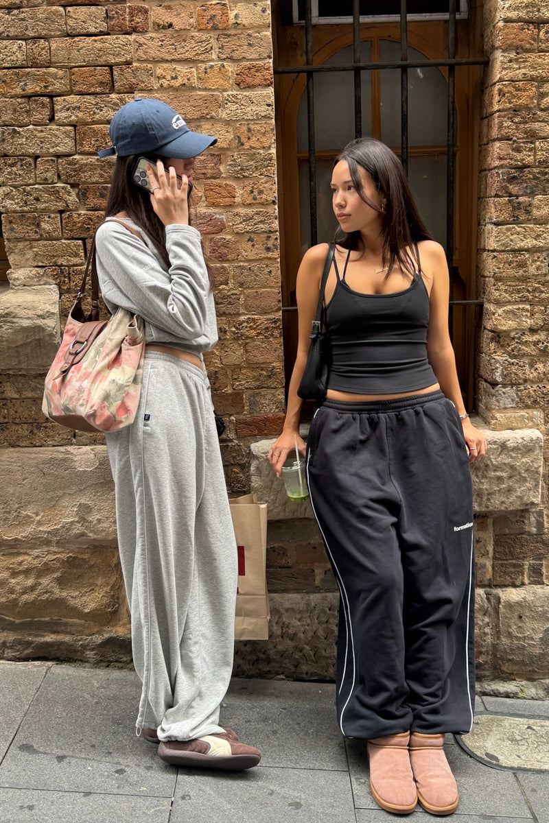 Two women standing against a brick wall, one in gray tracksuit and blue cap, the other in black outfit.