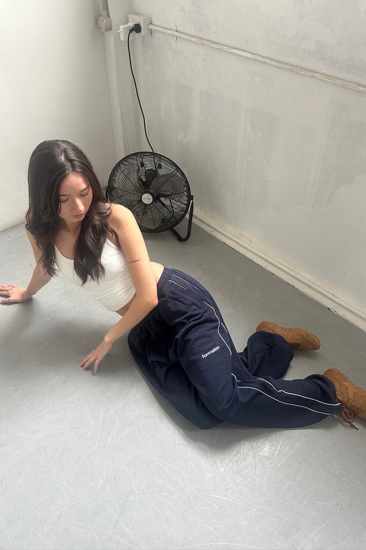 Woman sitting on the floor with a fan in the background. Wearing navy sweatpants and white tank top.