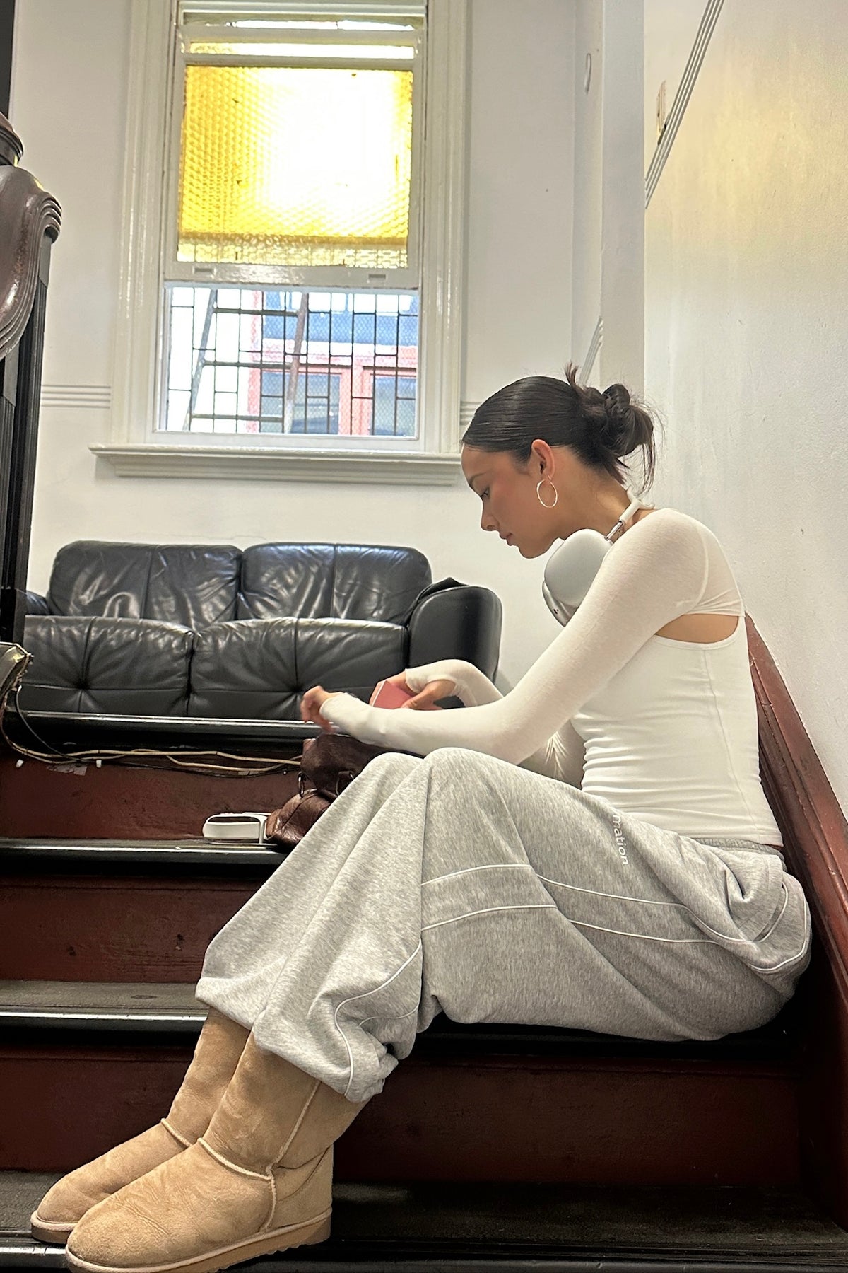 Woman sitting on a staircase wearing white bolero and tank top with grey sweatpants and beige UGG boots in a room with a couch and window.