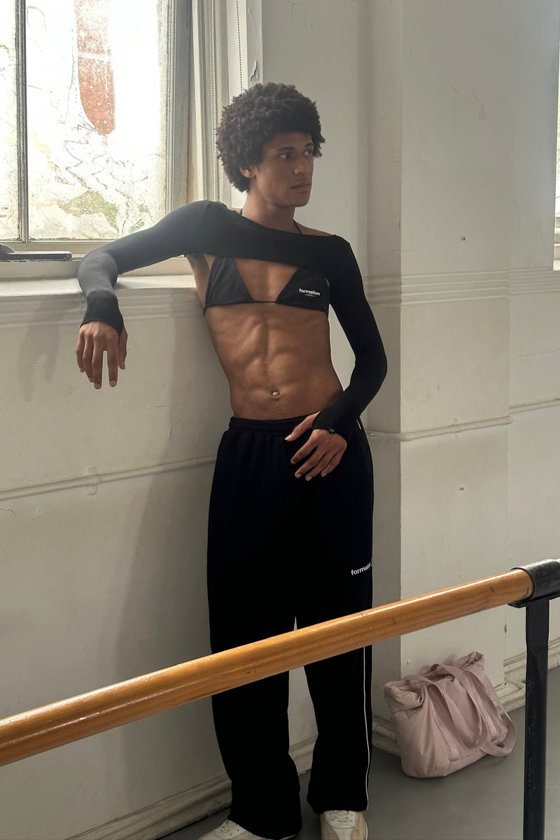 Person wearing a black bolero and sweatpants with triangle top, leaning against a wall with a window in the background.