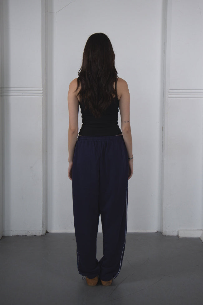 Person wearing a black sleeveless top and navy pants against a white wall.