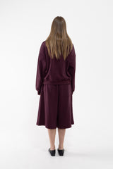 A dancer viewed from behind wearing a matching plum lounge set consisting of a long-sleeve sweatshirt and a knee-length skirt, paired with black slip-on shoes against a plain white studio background.