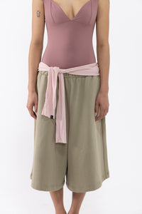A dancer wearing a dusty rose bodysuit, olive green wide-leg shorts, and a light pink fabric belt tied at the waist, shot against a plain white studio background.