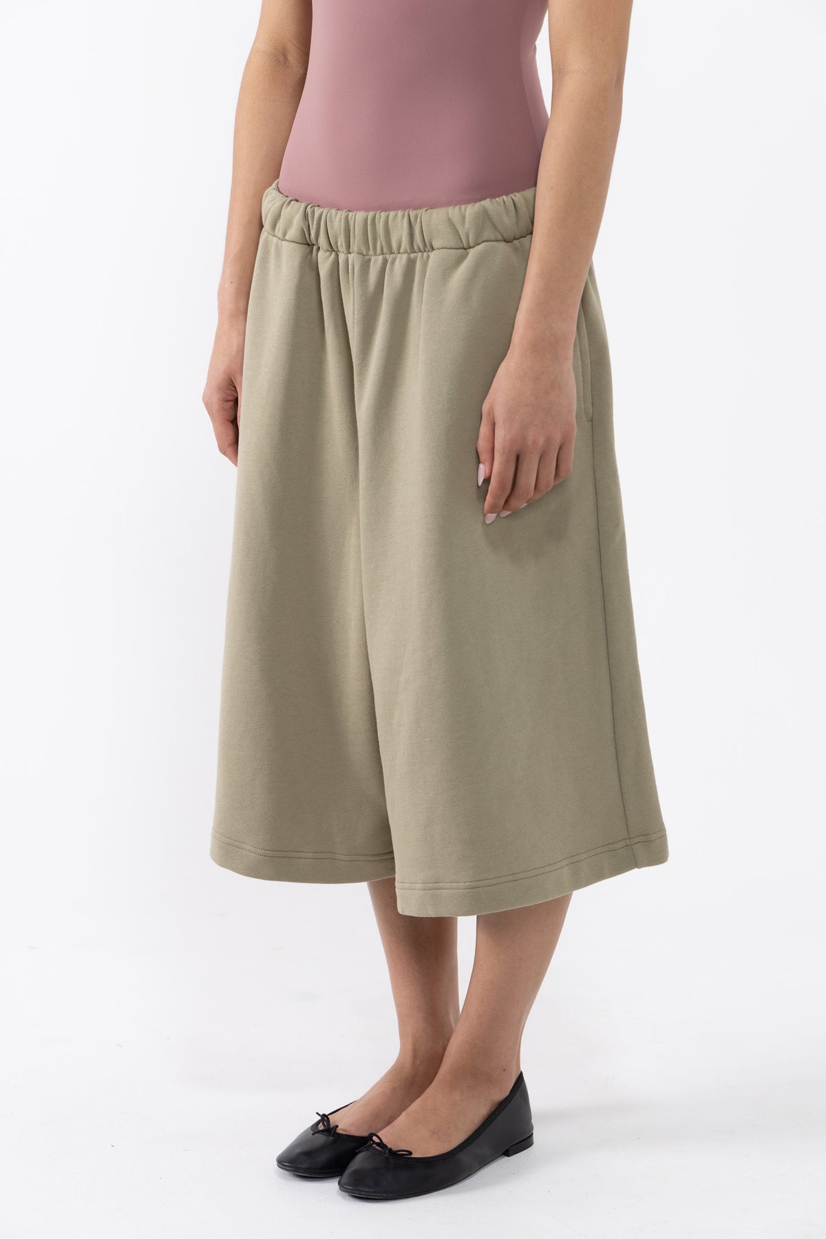 A dancer wearing a pair of wide-leg, knee-length olive green shorts with an elastic waistband, paired with a dusty pink top and black flats, against a plain white studio background.