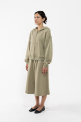 A dancer modeling a matching two-piece lounge set in a light sage green color, consisting of a zip-up hoodie and a midi skirt, against a plain white studio background.