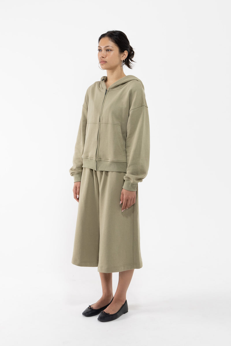 A dancer modeling a matching two-piece lounge set in a light sage green color, consisting of a zip-up hoodie and a midi skirt, against a plain white studio background.