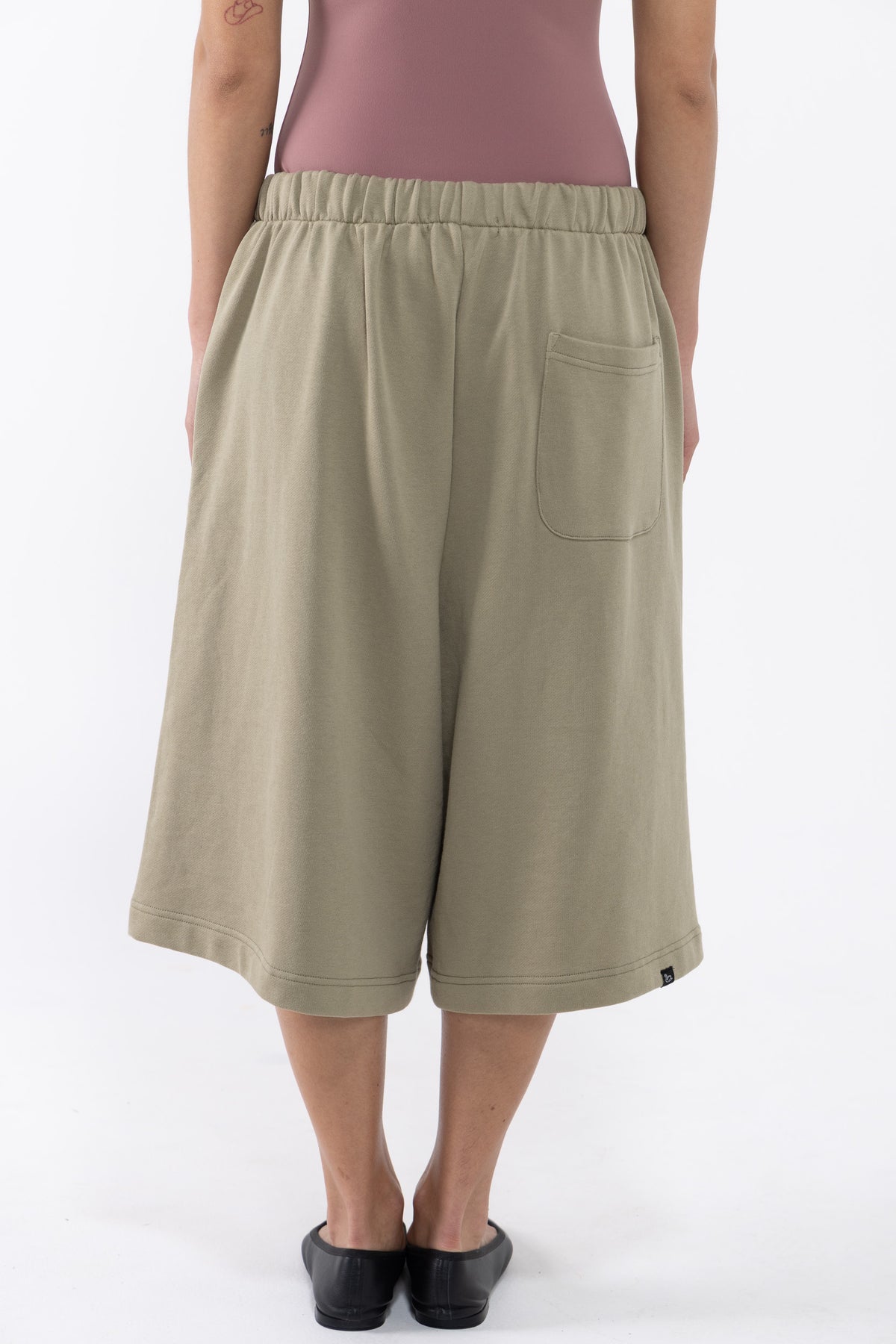 A dancer wearing loose, knee-length sage green shorts with an elastic waistband, a side pocket, and black slip-on shoes, shown against a plain light background.