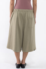 A dancer wearing loose, knee-length sage green shorts with an elastic waistband, a side pocket, and black slip-on shoes, shown against a plain light background.
