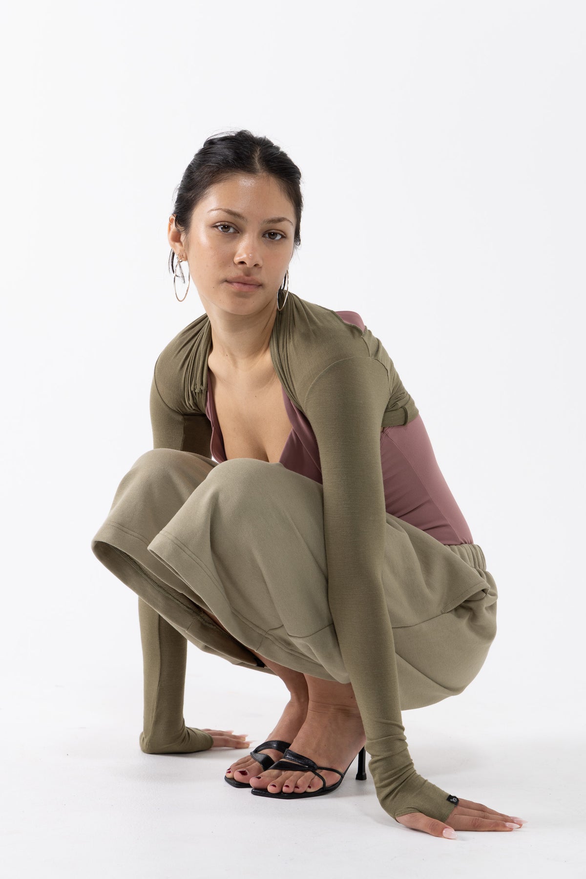 A dancer crouches in a studio setting wearing a coordinated olive green outfit consisting of a long-sleeve top, matching pants, and fingerless gloves, layered over a mauve-brown top, with black strappy heels.