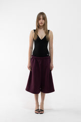 A dancer stands against a plain white studio background wearing a black sleeveless V-neck top and dark plum wide-leg shorts or culottes with black sandals.