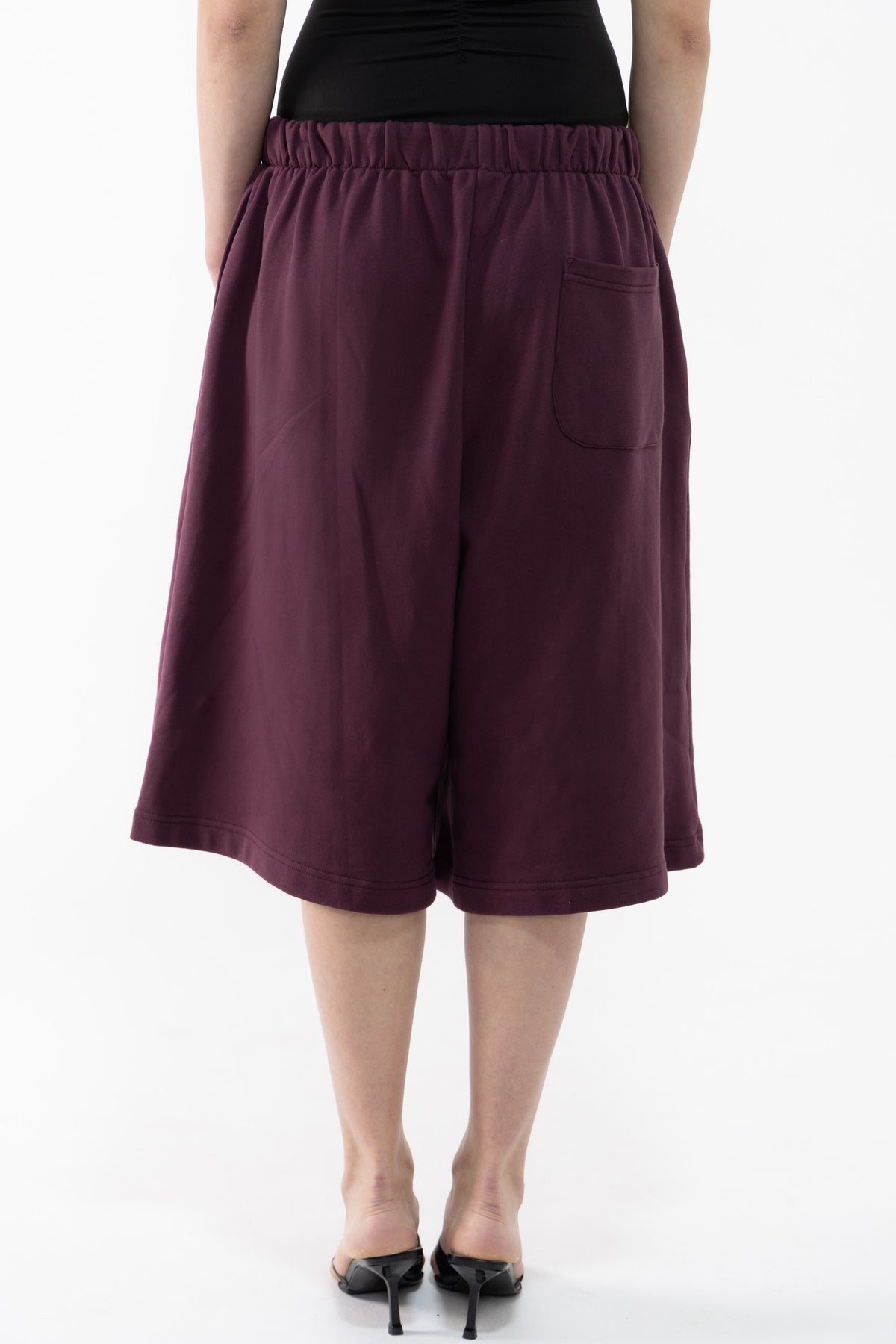 A dancer shown from the back wearing loose, wide-leg cropped shorts in a deep plum or burgundy tone, paired with a black top and black pointed heels, against a plain white studio background.