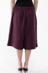 A dancer shown from the back wearing loose, wide-leg cropped shorts in a deep plum or burgundy tone, paired with a black top and black pointed heels, against a plain white studio background.