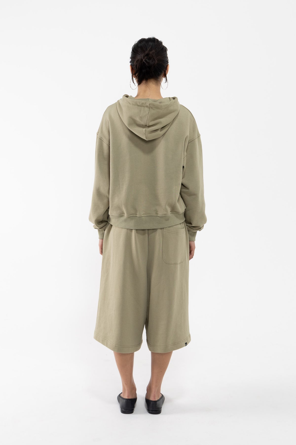 A dancer viewed from the back wearing a matching light olive green hooded sweatshirt and wide-leg shorts set, paired with black shoes, against a plain white studio background.