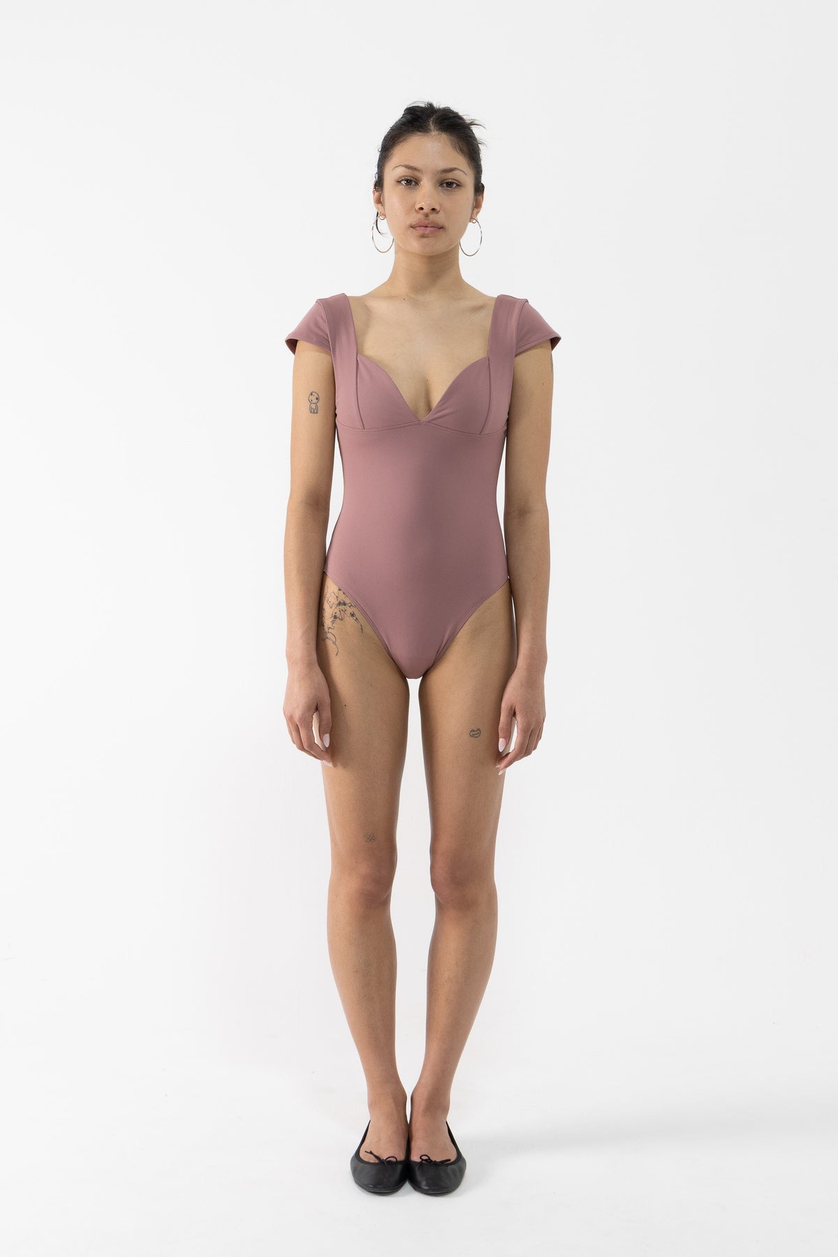 A dancer stands against a plain white studio background wearing a dusty rose one-piece leotard with short cap sleeves, a deep V-neckline, and side cutouts, paired with black flats.