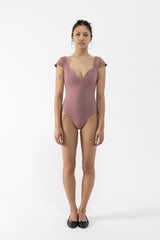 A dancer stands against a plain white studio background wearing a dusty rose one-piece leotard with short cap sleeves, a deep V-neckline, and side cutouts, paired with black flats.