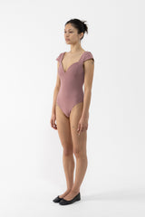 A dancer modeling a dusty rose pink one-piece leotard with cap sleeves and a deep V-neckline, standing in profile against a plain white studio background.