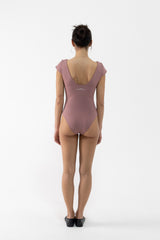 A dancer viewed from behind wearing a dusty mauve one-piece leotard with short cap sleeves and a deep V-back, paired with black ballet shoes, against a plain white studio background.