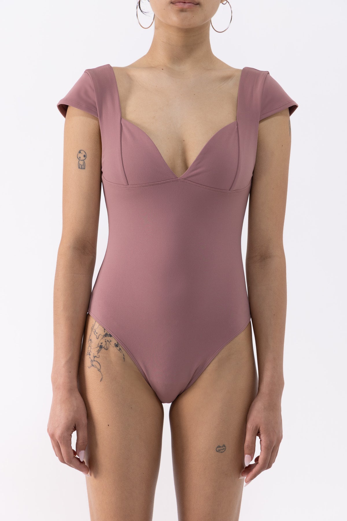 A dancer wearing a dusty rose one-piece leotard with cap sleeves and a deep V-neckline, shown from the neck down against a plain white background.