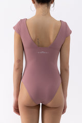 A dancer shown from the back wearing a dusty rose one-piece leotard with short cap sleeves, a deep V-neckline, and a small white decorative graphic across the upper back.
