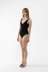 A dancer modeling a black one-piece leotard with a V-neckline and shoulder straps, standing against a plain white studio background.