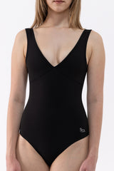 A dancer wearing a black one-piece leotard with a deep V-neckline and wide shoulder straps, shown against a plain light gray studio background.