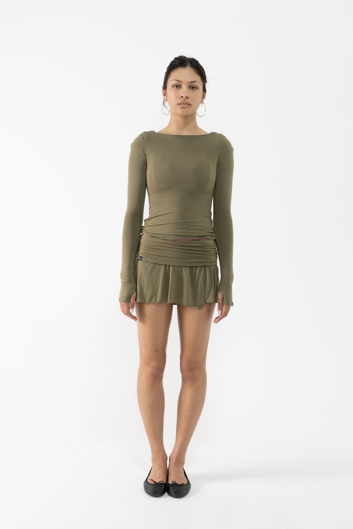 A dancer stands against a plain white studio background wearing a fitted long-sleeve top and mini skirt in olive green with ruching at the waist and black flats.