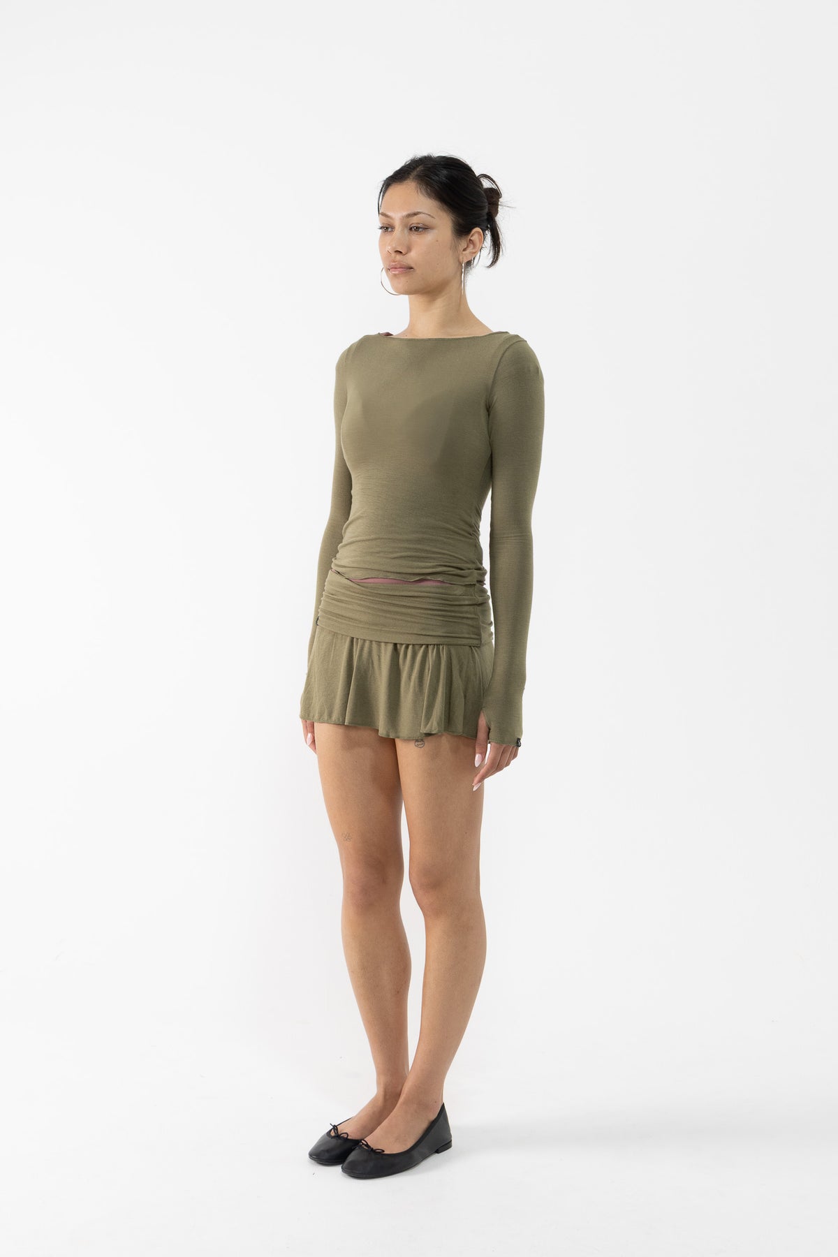 A dancer modeling a fitted, olive green long-sleeve top and mini skirt with ruching at the waist, standing against a plain white studio background.