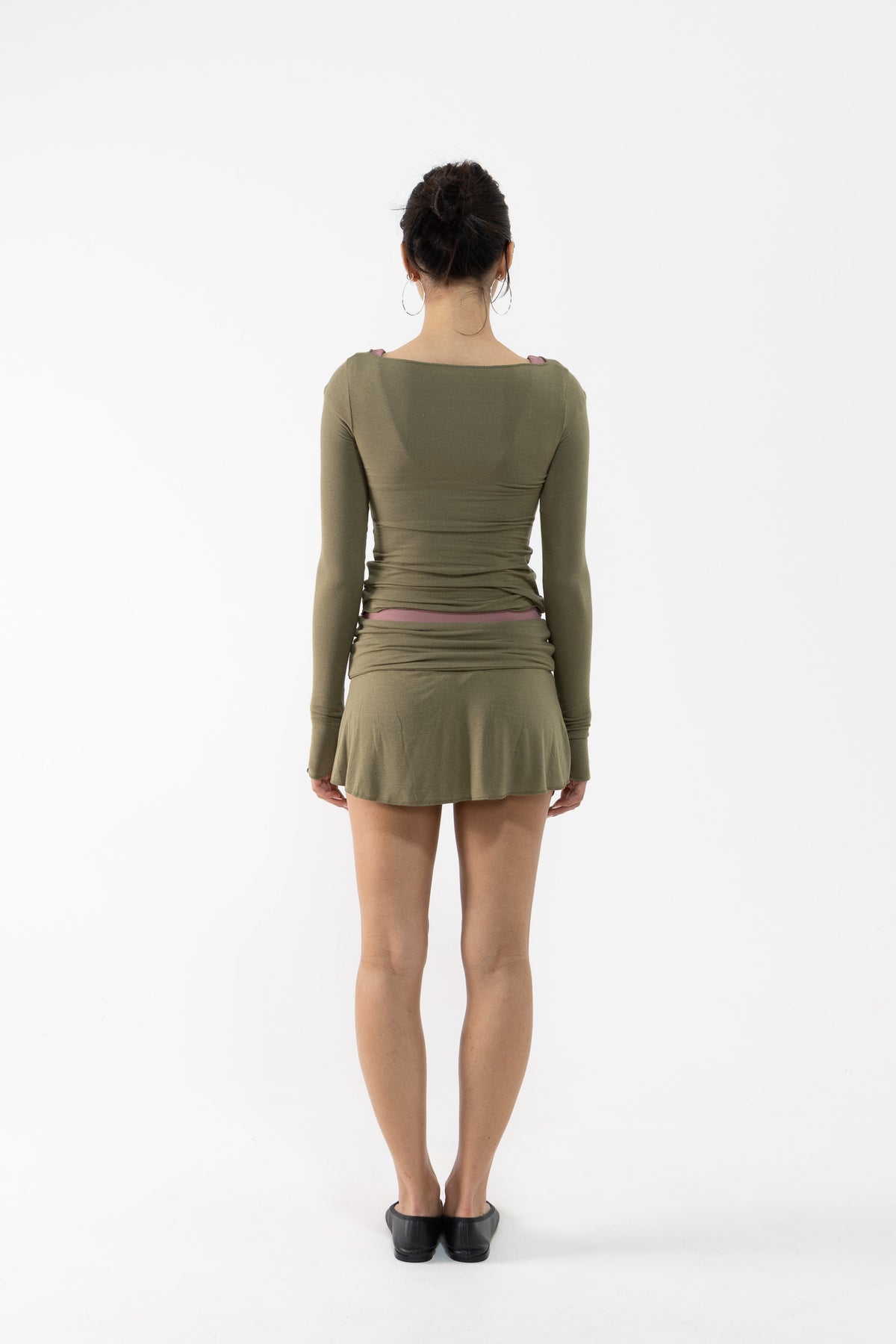 A dancer viewed from the back wearing a fitted long-sleeve top and mini skirt in olive green with ruching and a small pink waistband detail, standing against a plain white studio background.
