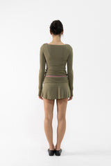 A dancer viewed from the back wearing a fitted long-sleeve top and mini skirt in olive green with ruching and a small pink waistband detail, standing against a plain white studio background.