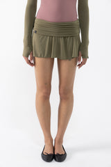 A dancer wearing an olive green ruched mini skirt with a matching long-sleeve top and black ballet flats, shown from the waist down against a white studio background.
