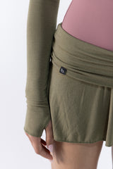 Close-up of a dancer wearing an olive green long-sleeve athletic wrap skirt or shorts with a pink top visible underneath, against a clean white background.