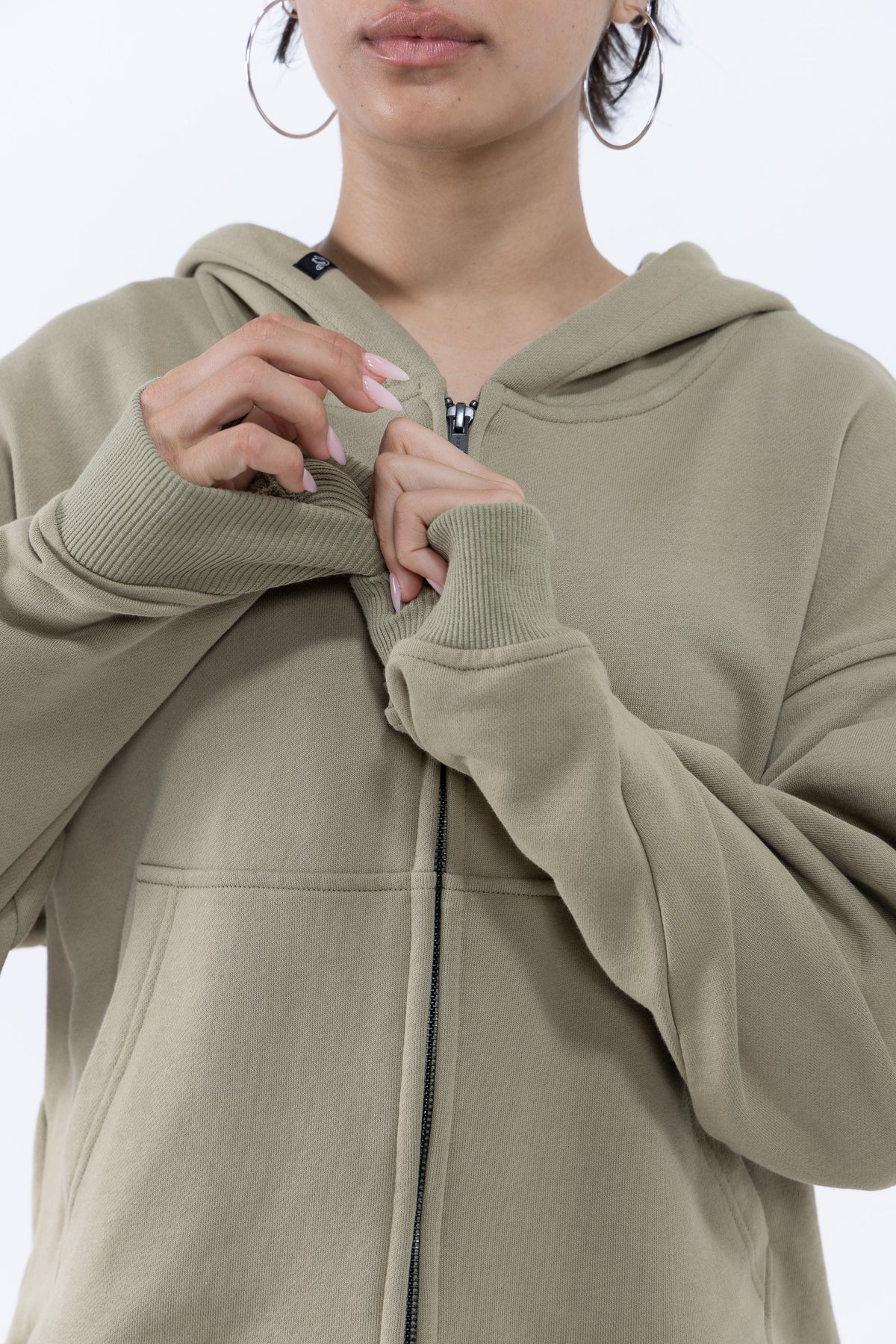 A close-up of a dancer wearing an olive green zip-up hoodie, adjusting the zipper. The person is also wearing large silver hoop earrings, set against a plain white background.