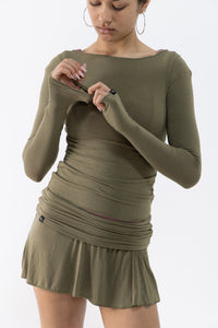 A dancer wearing an olive green long-sleeve ruched top and mini skirt with a boat neckline, shown from the head down against a plain white background.