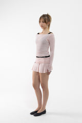 A dancer modeling a light pink long-sleeve athletic top and matching pleated mini skirt with a thin black waistband, paired with black flats, against a plain white studio background.
