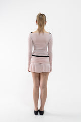A dancer stands with her back to the camera wearing a light pink long-sleeve mini dress with black trim details, set against a plain white studio background.