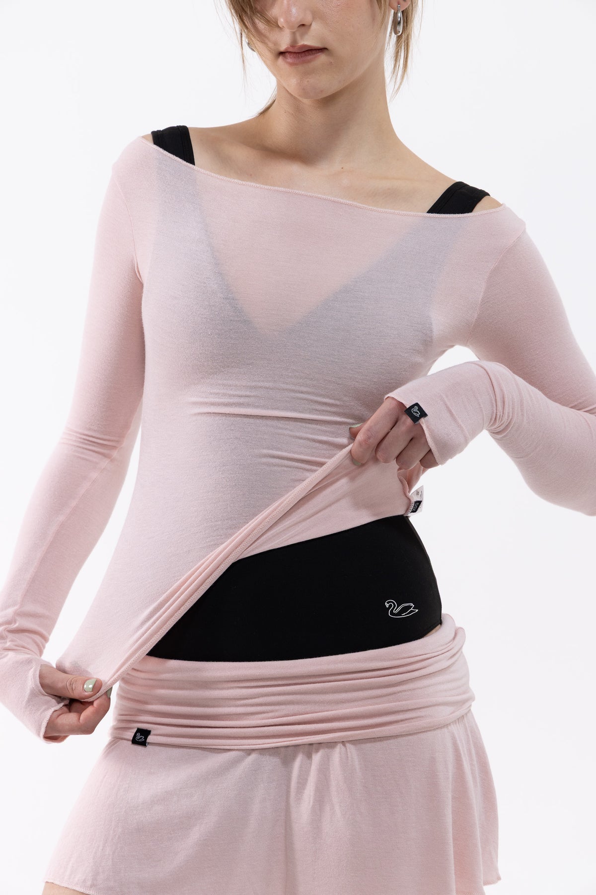 A dancer wearing a light pink long-sleeve athletic top and matching skirt, lifting the top to reveal a black waist support band with a small white logo.