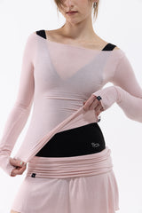 A dancer wearing a light pink long-sleeve athletic top and matching skirt, lifting the top to reveal a black waist support band with a small white logo.
