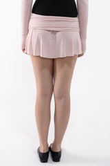 Rear view of a model wearing a light pink ruched wrap mini skirt with long sleeves, paired with a black top and black shoes, on a white studio background.