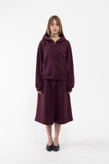 A dancer stands against a plain white studio background wearing a matching plum zip-up hoodie and wide-leg cropped sweatpants, paired with black flats.
