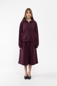 A dancer stands against a plain white studio background wearing a matching plum zip-up hoodie and wide-leg cropped sweatpants, paired with black flats.