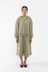 A dancer stands against a plain white studio background wearing a matching sage green two-piece lounge set consisting of a zip-up hooded sweatshirt and wide-leg cropped pants.